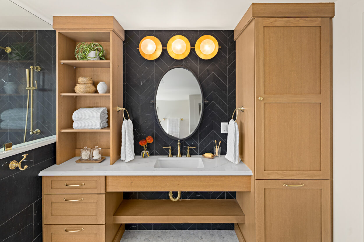 Gold-framed pendant lights on this vanity become a visual focal point in this black and wood-toned bathroom. 2025 design trends list statement lighting to create visual interest, so these bulbs get the job done. 